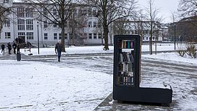 Der Bücherschrank befindet sich rechts im Bild. An ihm vorbeischauen sieht man die Häuser 1 und 18. Über allem liegt eine dünne Schneedecke.