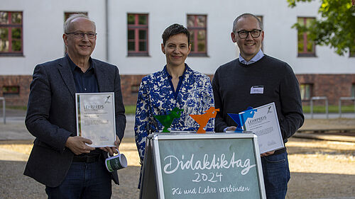 Die drei Personen stehen nebeneinander hinter einem Aufsteller mit dem Titel "Didaktiktag 2024". Sie halten ihre Urkunden in der Hand. Auf dem Aufsteller stehen die drei Preispokale in Form des Hochschulfischers.