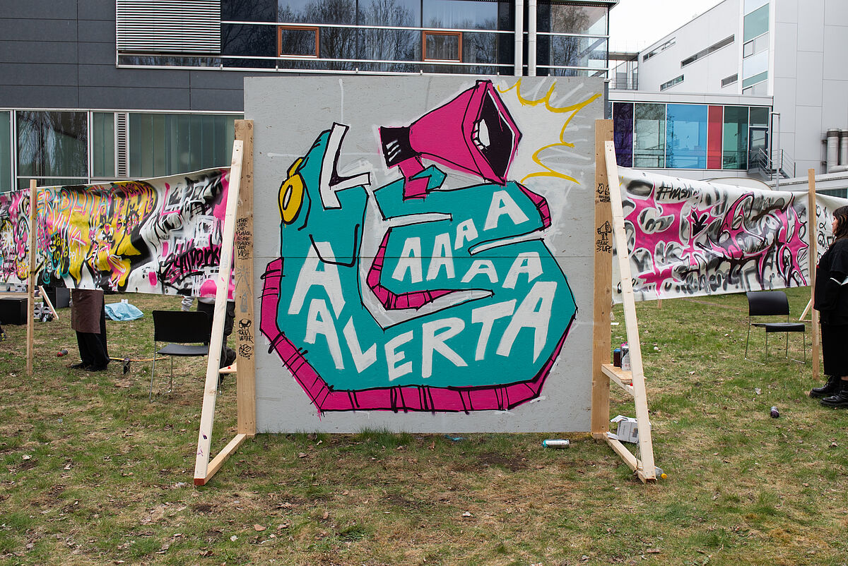 Eine besprayte Wand aus dem Graffiti-Workshop mit farbenfrohen Motiven, darunter ein Fisch mit Megafon und dem Schriftzug „Alertaaaaa“. Die Gestaltung spiegelt kreative Auseinandersetzungen mit dem Thema Inklusion wider.